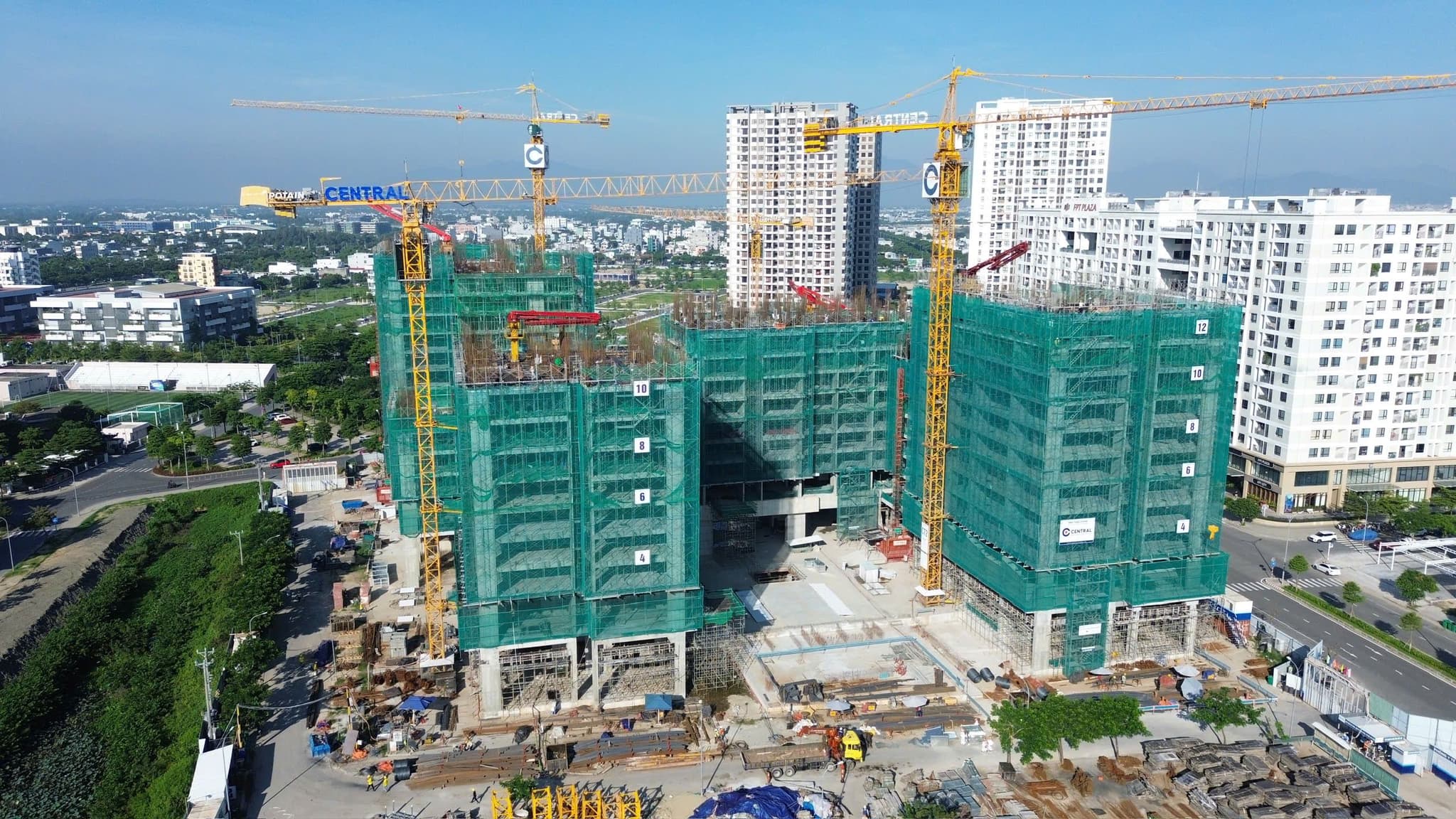 Aerial view of a high-rise construction site with tower cranes, green safety netting, and urban apartments.