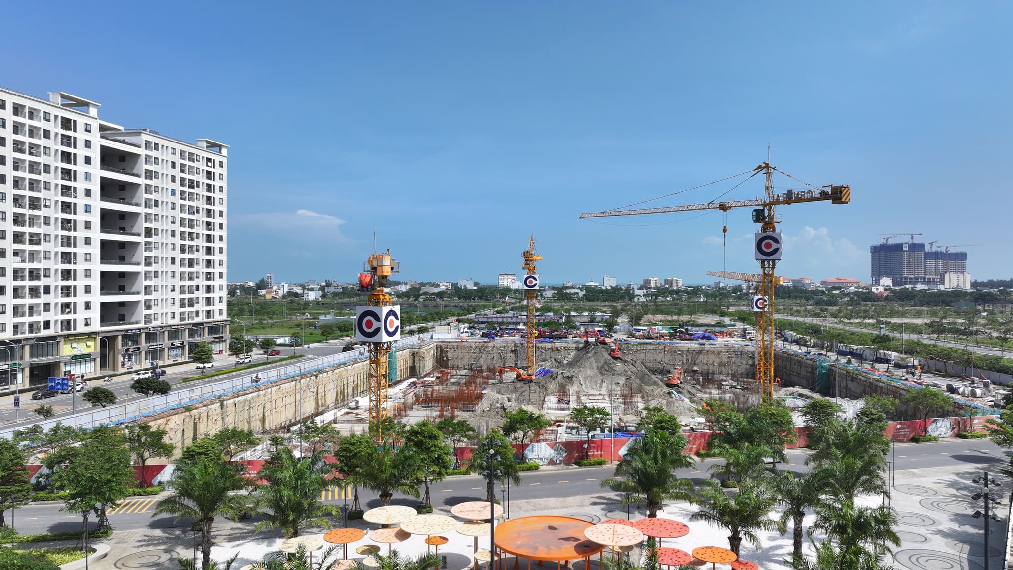 Urban construction site with tower cranes beside high-rise apartments and a palm-lined plaza under blue sky