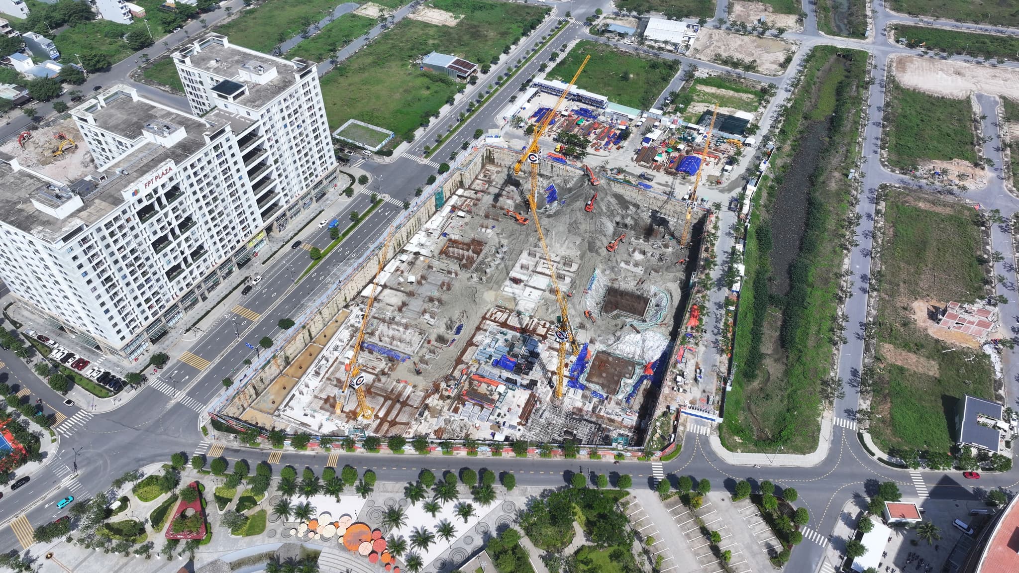 Aerial view of a large urban construction site with tower cranes, apartment buildings, and tree-lined roads